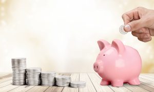 Man putting coin in pig moneybox on table with coin stacks